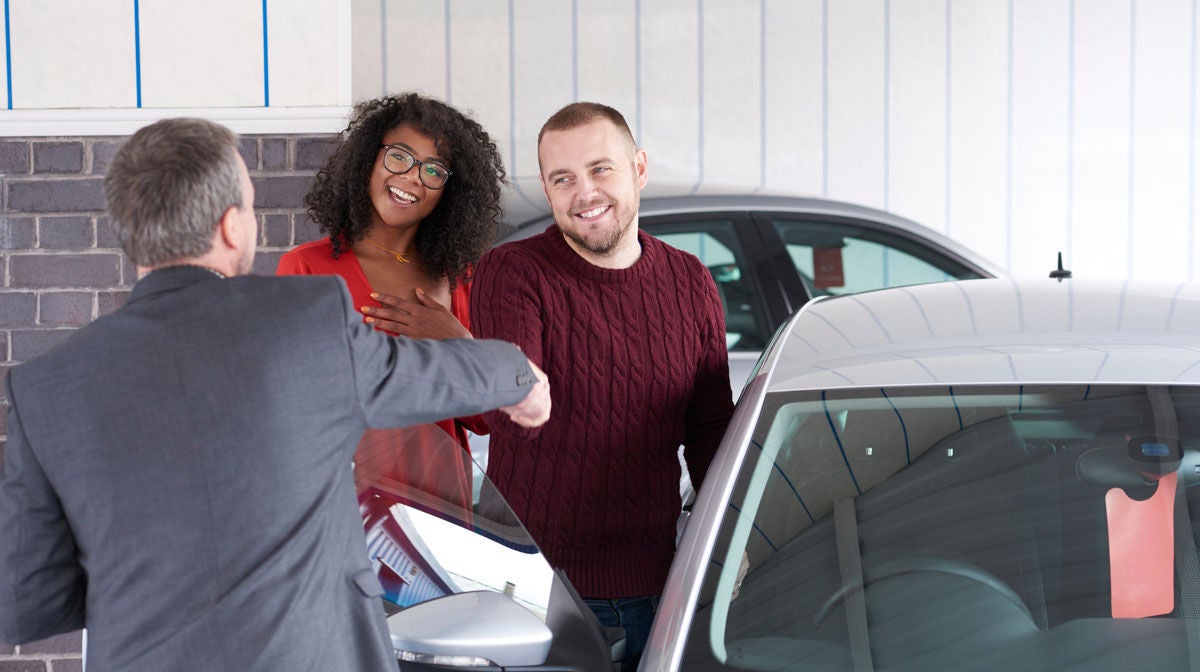 A young couple are looking at  cars in a car showrooms . A salesman is explaining about the car whilst the young couple listen and look at a car .