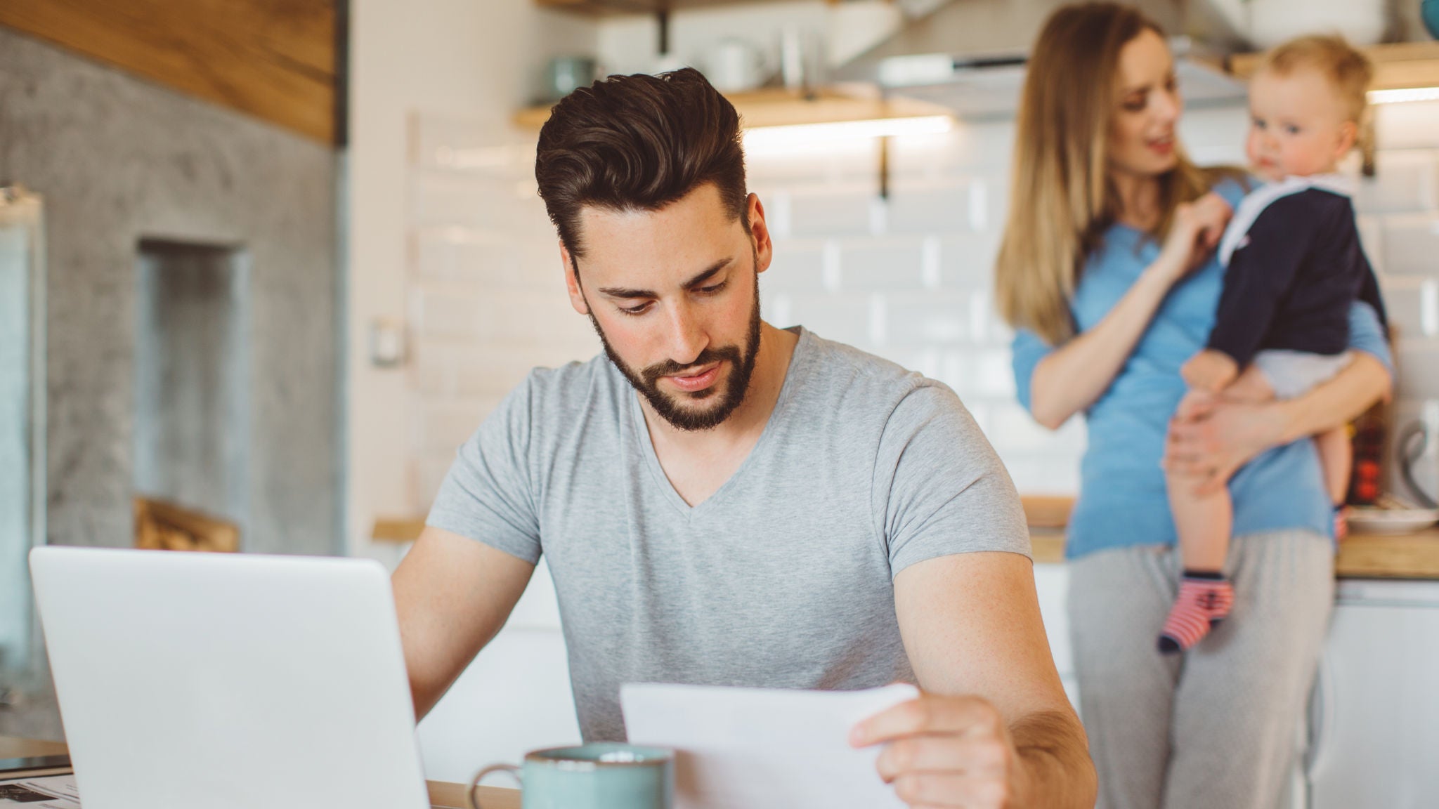 Young couple considering refinancing at home with their baby boy. They are wearing pajamas and drinking first coffee of the day at the kitchen.