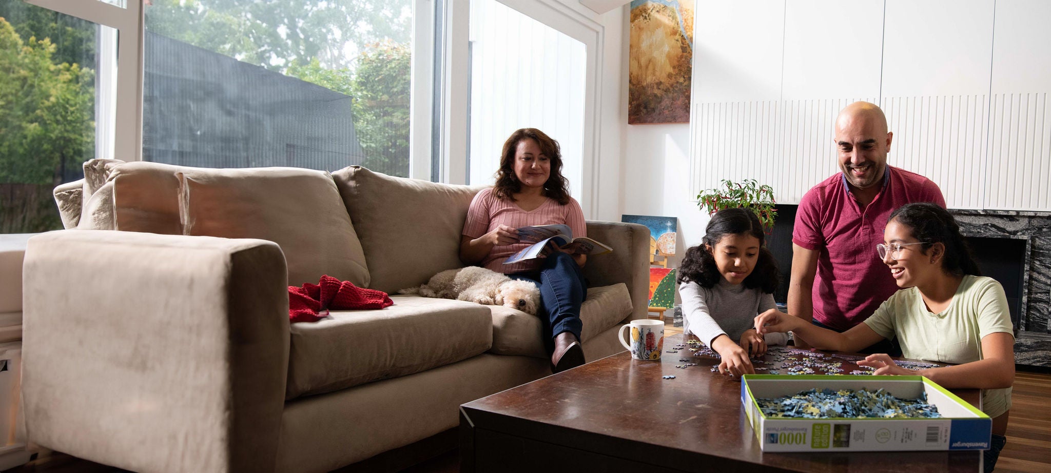 Family assembling a puzzle at a table while a dog rests on a couch in a living room